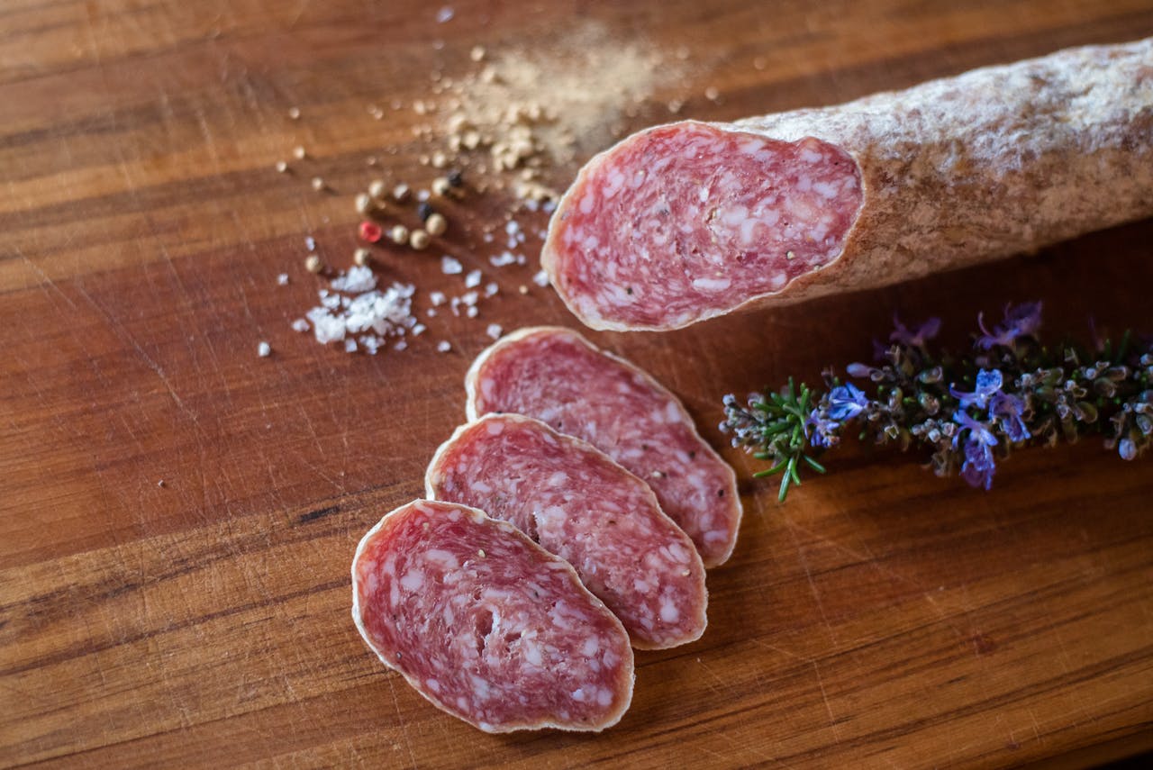 Sliced salami arranged on a wooden board with whole peppercorns, sea salt, and fresh rosemary flowers