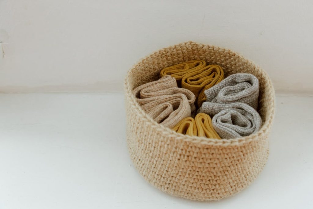 round woven basket filled with neatly rolled beige, mustard, and gray fabric items on white surface near wall