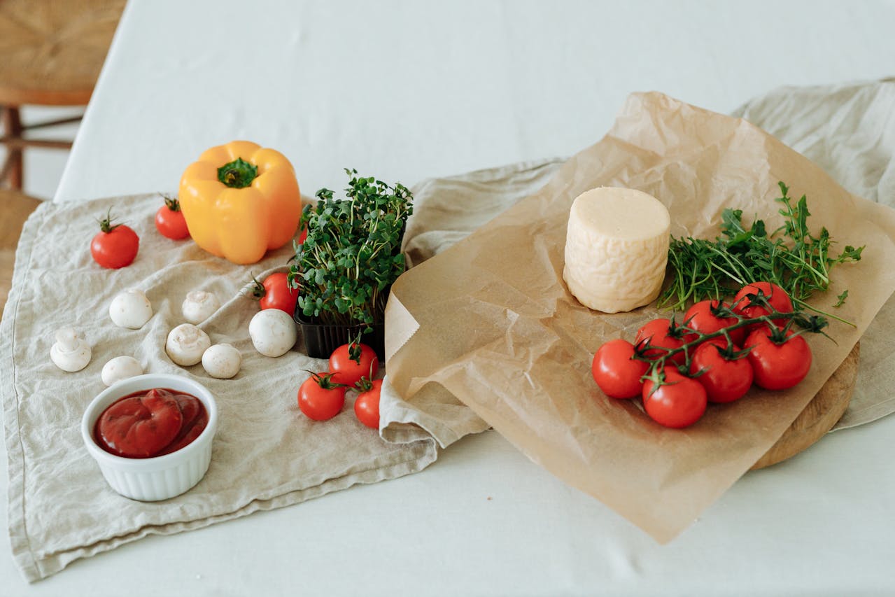 Fresh vegetables and cheese on a table, including cherry tomatoes, yellow bell pepper, mushrooms, greens, and soft cheese