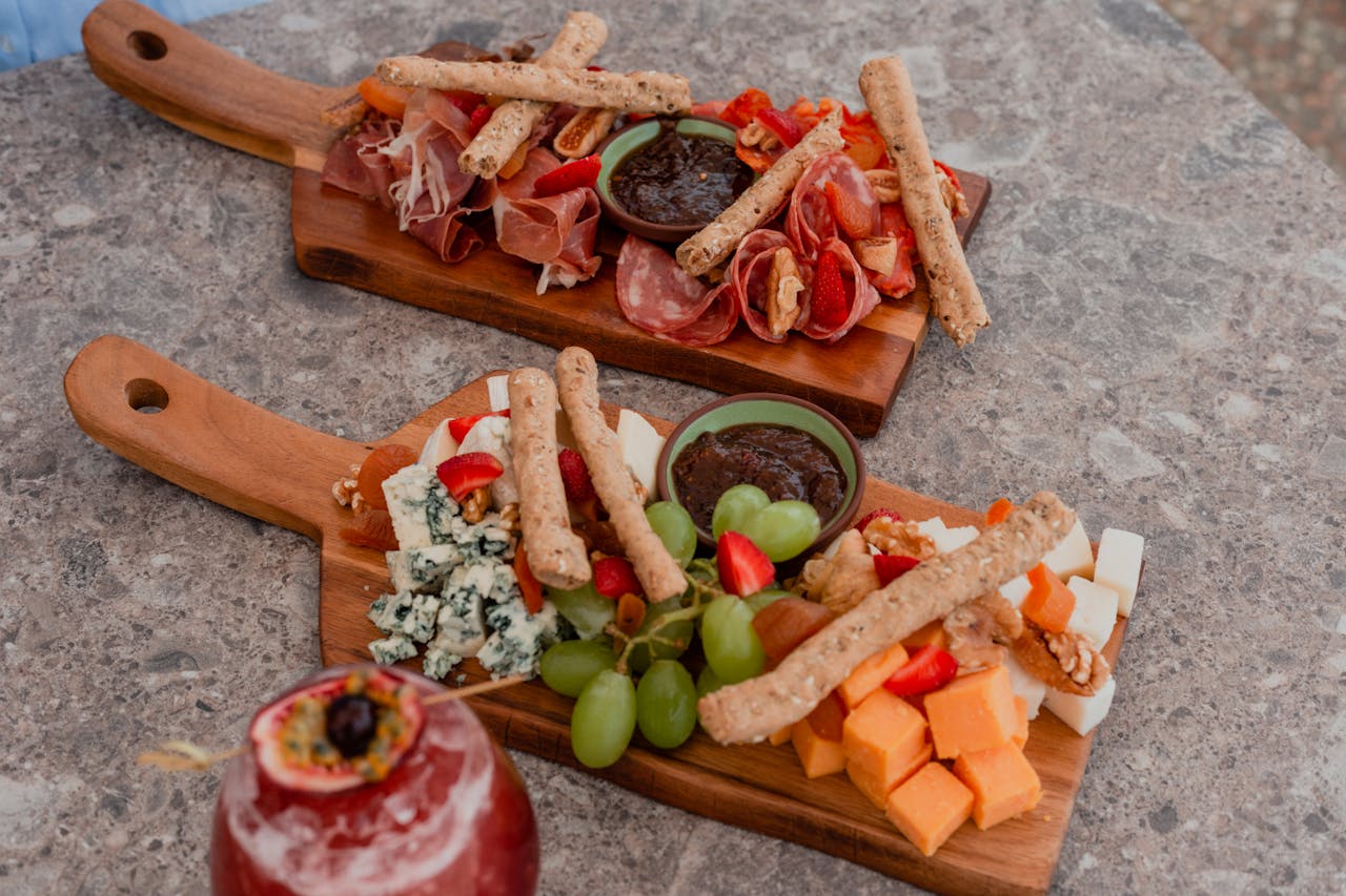 Two colorful charcuterie boards with assorted cheeses, meats, grapes, strawberries, jam, and breadsticks