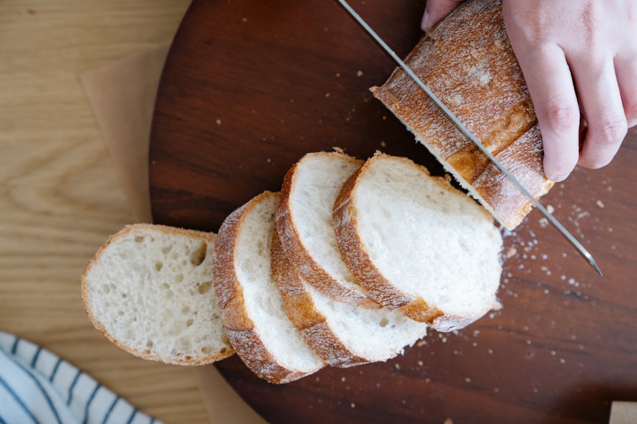 Freshly sliced baguette on a wooden board, hand holding and cutting loaf with a bread knife