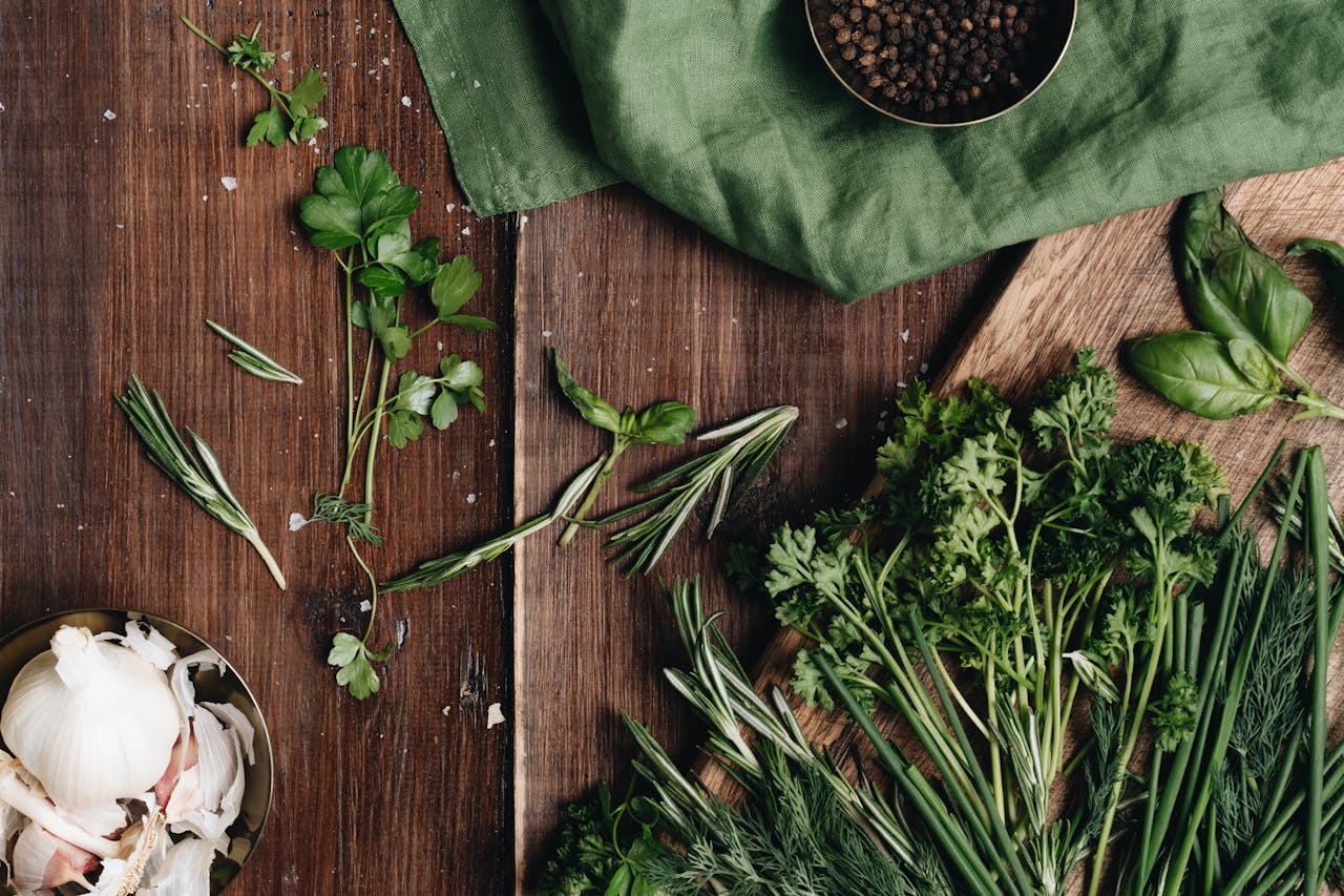 Fresh herbs including rosemary, parsley, basil, and dill scattered on a wooden surface with garlic and peppercorns