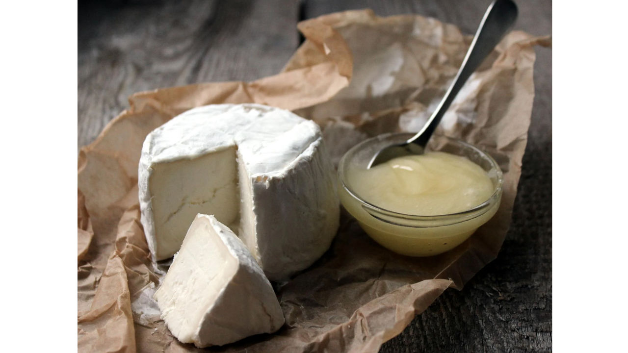 Wheel of soft-ripened goat cheese with a wedge cut out, served on brown parchment with a small glass bowl of honey and spoon beside it