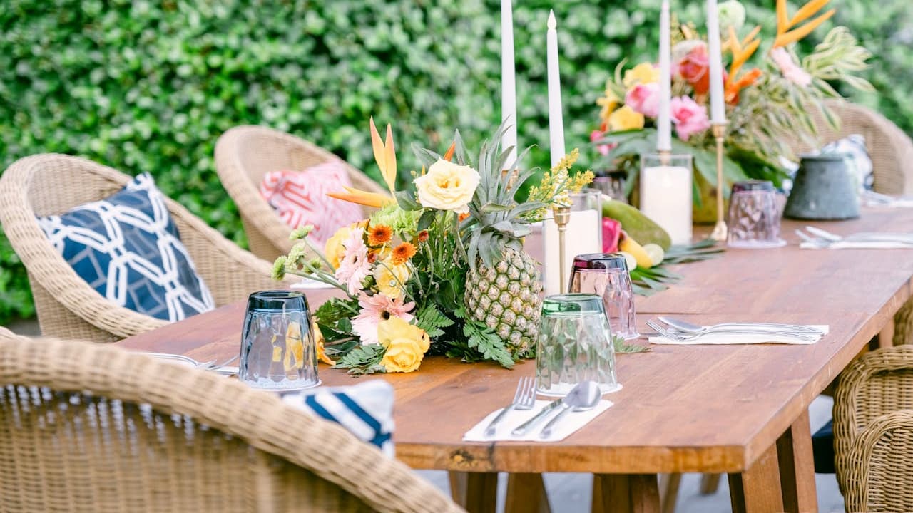 Outdoor wooden dining table set with floral centerpieces, tropical fruits, glassware, candles, and wicker chairs with decorative cushions, lush green hedge in the background, bright and festive setup