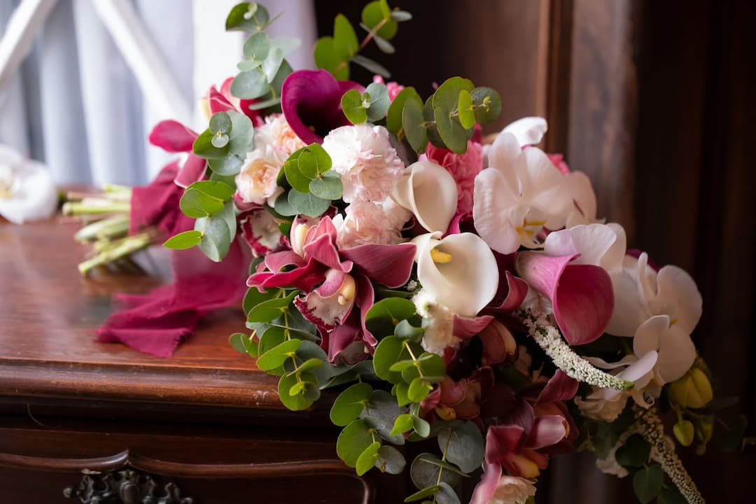 elegant bouquet of pink peonies, white and burgundy calla lilies, orchids, roses, and eucalyptus leaves, tied with a burgundy ribbon, resting on a polished wooden surface