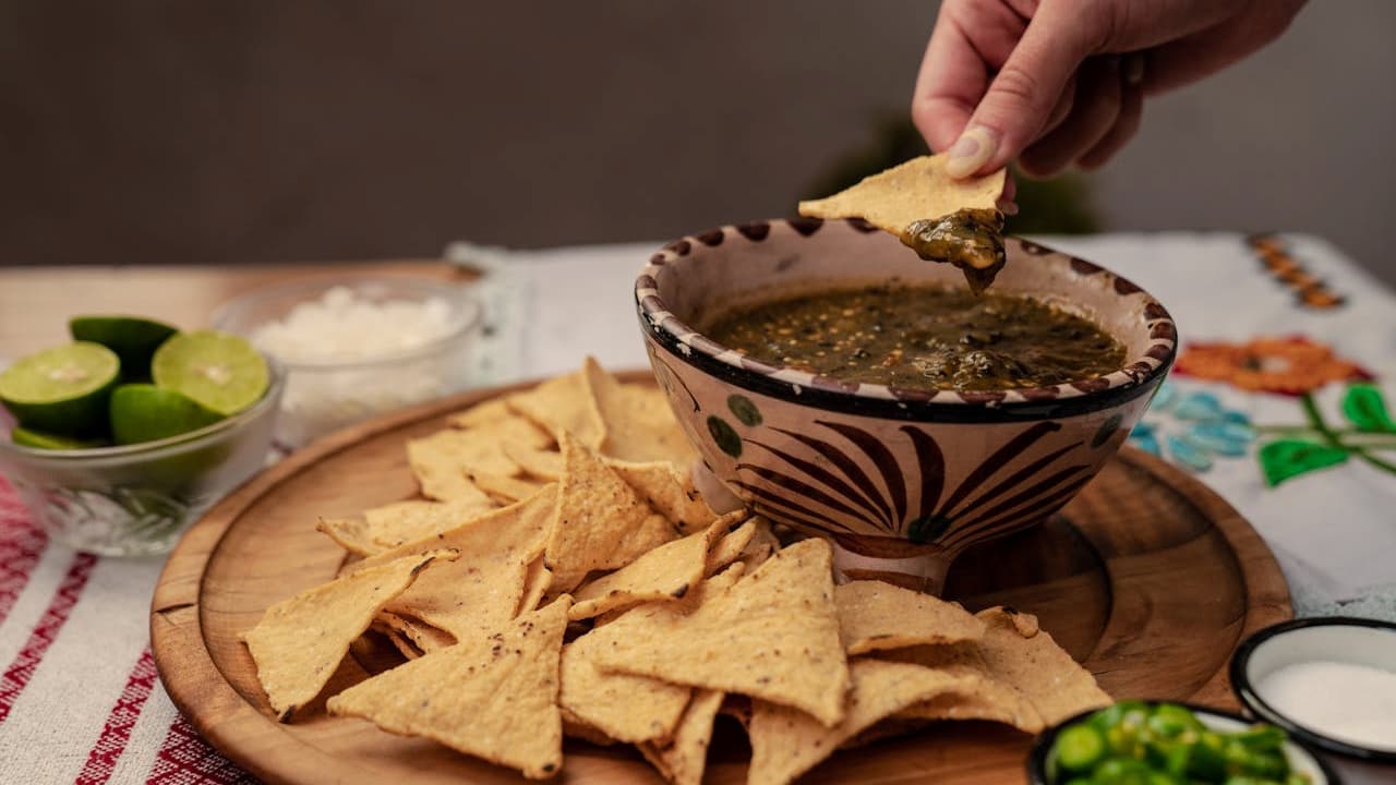 Hand dipping a tortilla chip into a bowl of green salsa, wooden platter filled with crispy tortilla chips, garnished with lime slices, chopped green chilies, and sour cream in the background, colorful tablecloth underneath