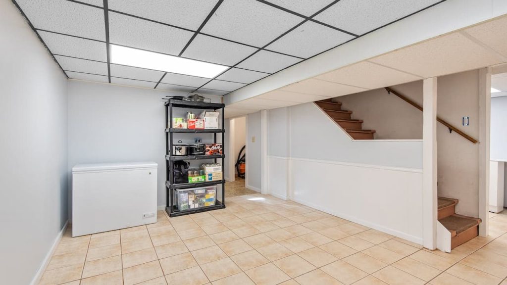 Basement room with tiled floor, white chest freezer on the left wall, black metal shelving unit stocked with household supplies, stairs with wooden steps leading up on the right, white walls and ceiling with grid-style fluorescent lighting
