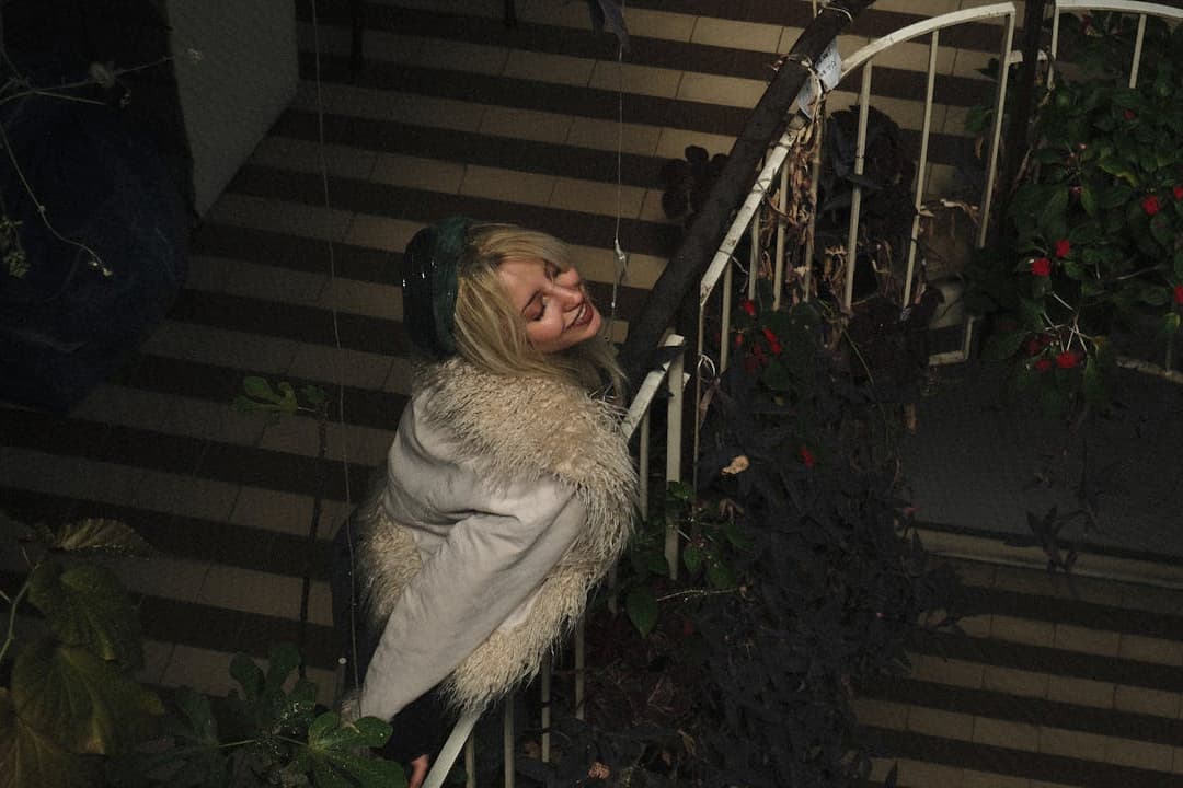 Blonde woman wearing a white coat with faux fur trim and a green headband, smiling with eyes closed, leaning back playfully on a spiral staircase, surrounded by potted plants with red flowers