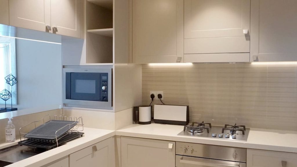 Modern kitchen interior, beige cabinetry with under-cabinet lighting, built-in microwave, four-burner gas stove with metallic knobs, tiled backsplash, window with natural light, cutting board and utensils holder on the counter, clean and minimalist design
