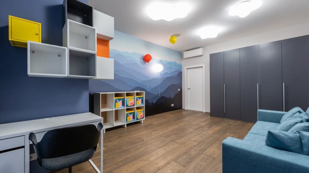 Modern, brightly lit kids’ room, blue and white theme, wall-mounted shelves with yellow and orange accents, study desk with black chair, mountain-themed wall mural, colorful bookshelf with toys and books, large gray wardrobes, teal sofa with matching cushions, wood flooring, ceiling lights, air conditioner above white door