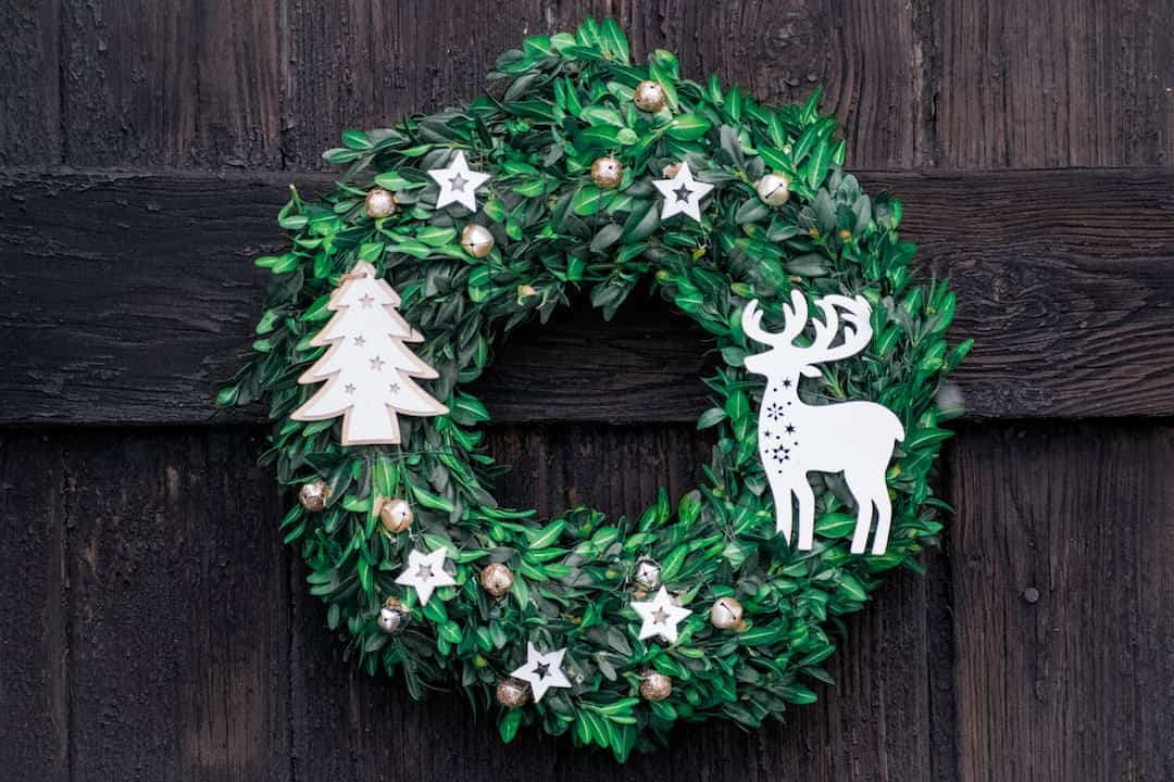 green Christmas wreath with gold baubles, white star ornaments, wooden tree and reindeer decorations, hanging on dark wooden door