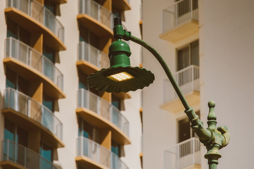 Vintage green street lamp with scalloped shade and LED light panel, set against a modern building with repeating balconies and sunlit exterior