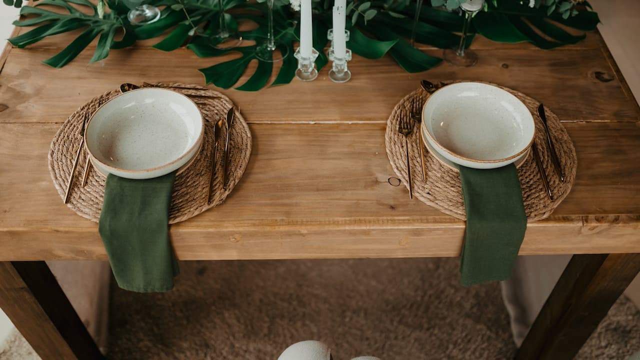 Minimalist table setting for two, wooden table, white bowls on woven placemats, green cloth napkins, leafy green centerpiece, soft natural lighting