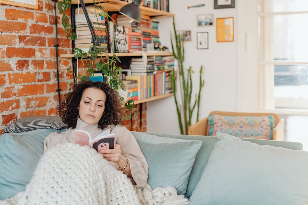 Woman relaxing on a blue couch, reading a book, wrapped in a chunky white blanket, surrounded by pillows, indoor plants, and a large bookshelf filled with books