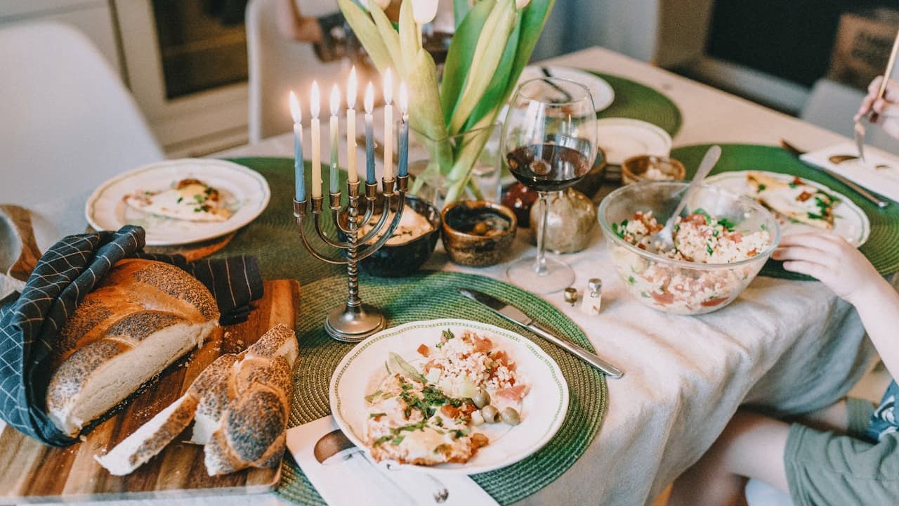 Casual dining table set with mismatched plates of food, green placemats, assorted breads, lit candles, leafy centerpiece, and a relaxed meal setting with people seated, warm and inviting atmosphere.
