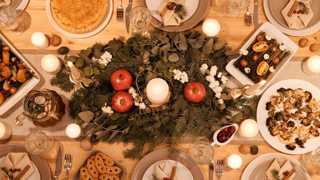 Festive dinner table setting, overhead view, decorated with candles, pine branches, red and white ornaments, surrounded by various dishes including roasted meats, vegetables, pasta, and desserts, warm lighting creating a cozy, celebratory atmosphere