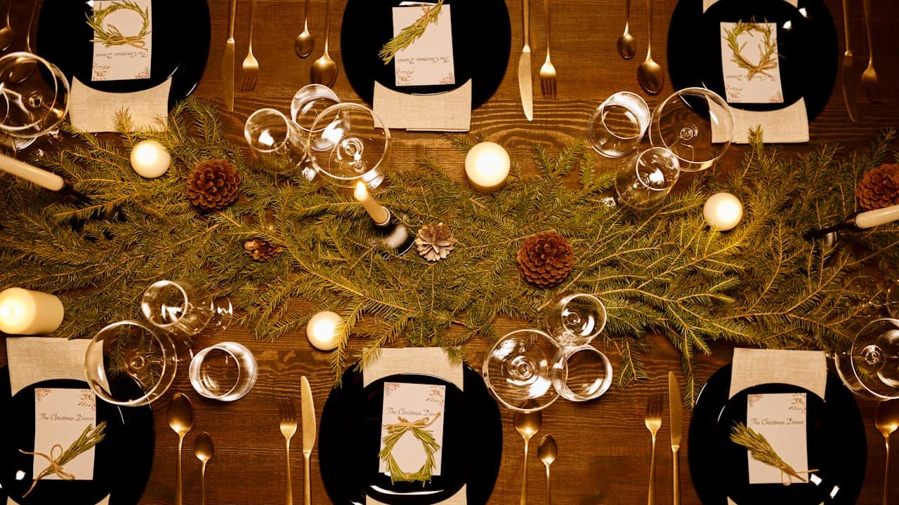 Elegant dinner table setting, overhead view, with black plates, gold cutlery, white napkins with gold rings, multiple wine glasses, candles, and a centerpiece of pine branches and pinecones, warm ambient lighting