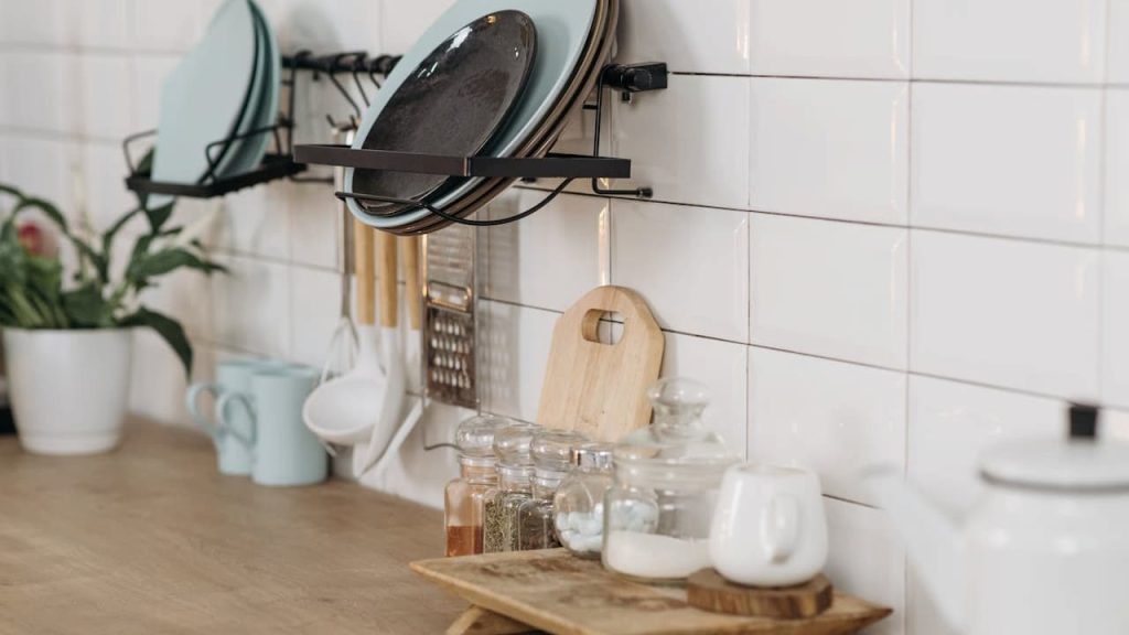 Neatly organized kitchen counter, white tiled backsplash, hanging rack with blue and black plates, wooden utensils hanging below, potted plant on the left, mugs and spice jars on the counter, small tray with a teapot and cup