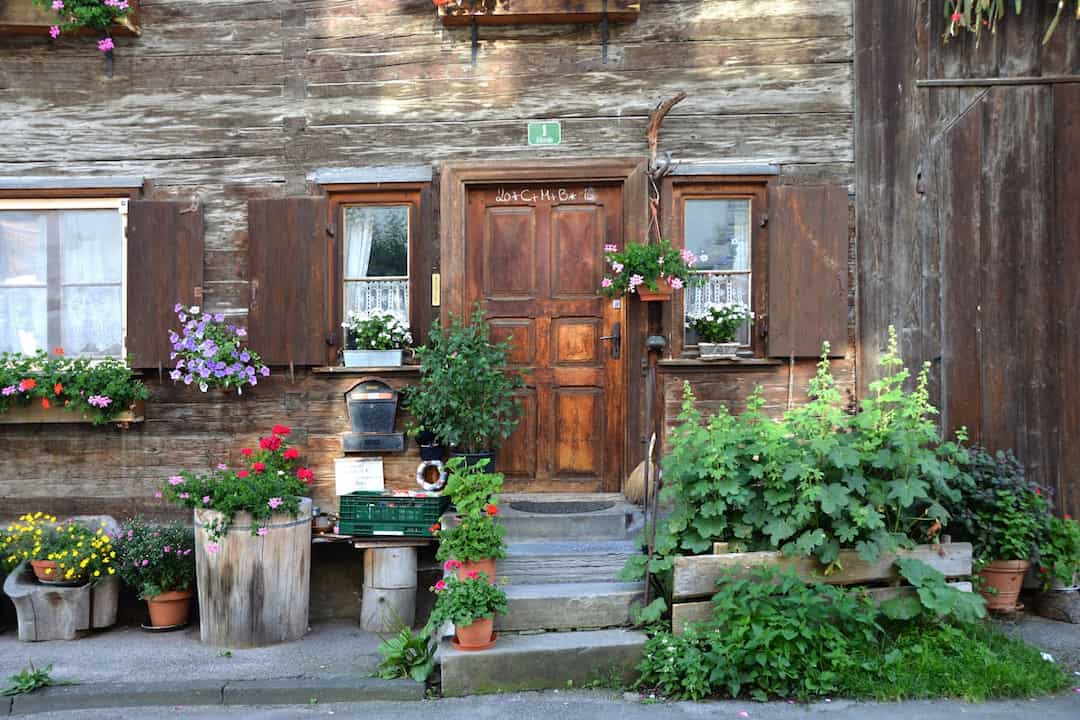 rustic wooden house with flower-filled window boxes and hanging baskets, wooden door with small steps, potted plants and greenery arranged around entrance