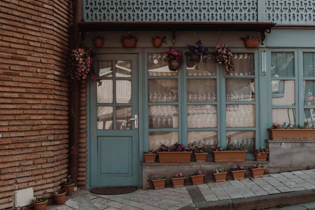 vintage-style building with blue-framed glass door and windows, lace curtains, flower pots on windowsills and sidewalk, hanging and wall-mounted planters, brick wall on the left