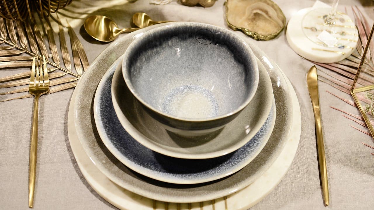 Elegant table setting with stacked ceramic dishes, blue and white gradient bowls, gold cutlery arranged on both sides, decorative coasters, white tablecloth with golden leaf accents, sophisticated and luxurious dining presentation