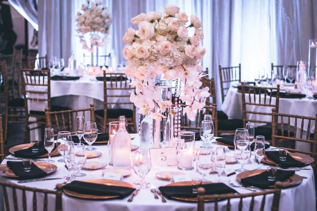 round wedding reception table with elegant place settings, glassware, and a tall floral centerpiece of white roses, hydrangeas, and cascading orchids, surrounded by gold chiavari chairs