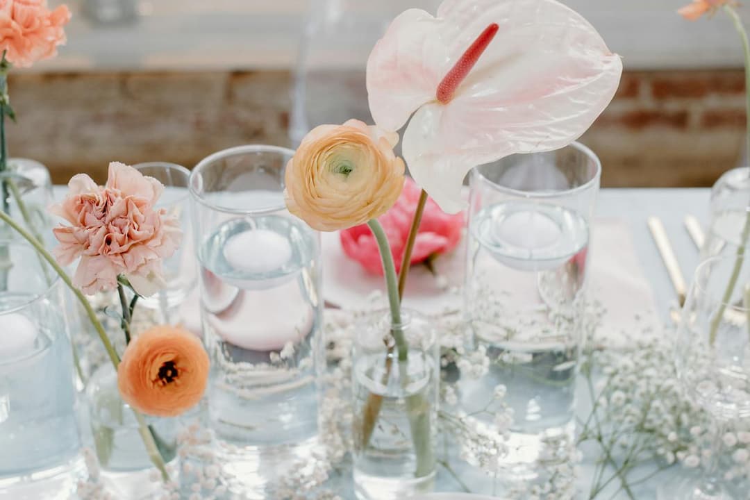 Close-up of a table centerpiece featuring a peach ranunculus, pink carnation, and pale pink anthurium in glass vases, surrounded by water-filled glasses and delicate white baby's breath