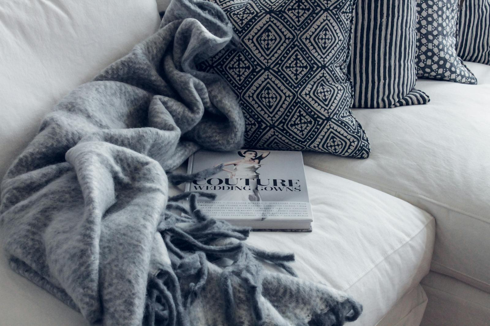 A stylish living room featuring a white sofa with patterned pillows, a blanket, and a fashion book.