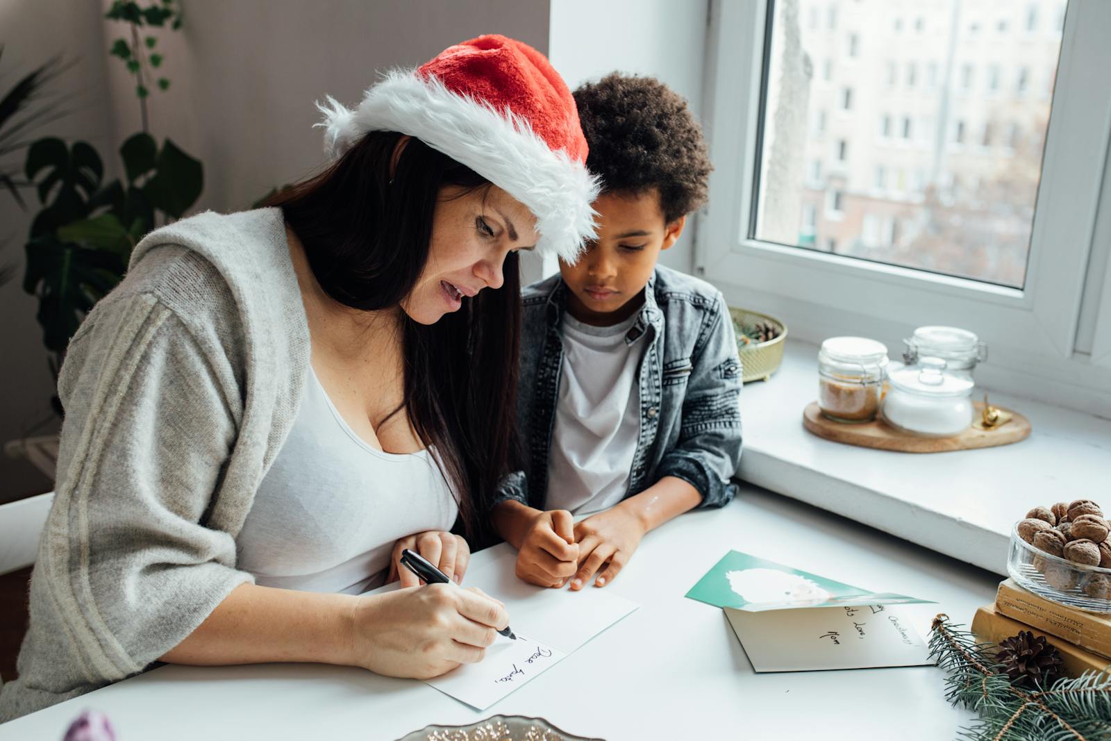 A mother and her son enjoy a festive moment writing Christmas cards by a window.