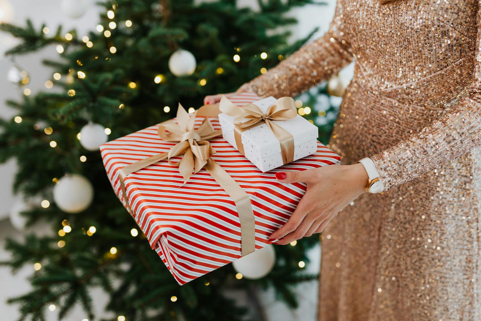 A woman in a sparkling dress holds beautifully wrapped gifts near a decorated Christmas tree.