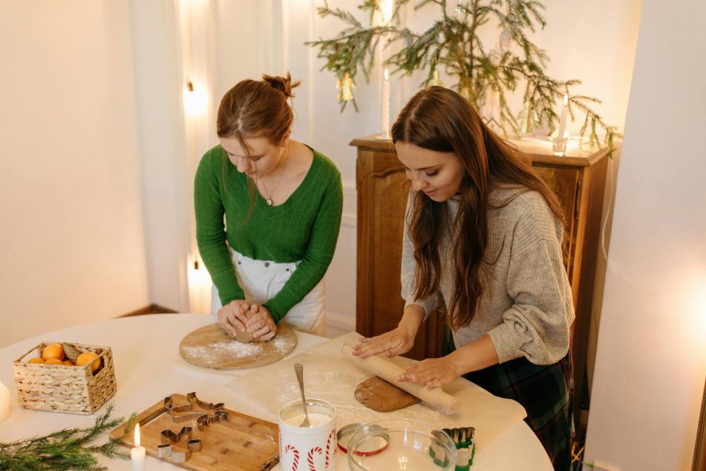 Two women enjoy Christmas baking indoors, kneading dough near a decorated spruce.
