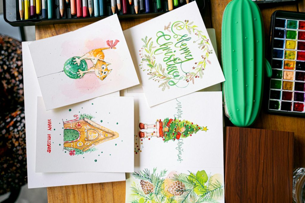 Top view of handmade Christmas cards with artistic designs on a wooden table surrounded by art supplies.