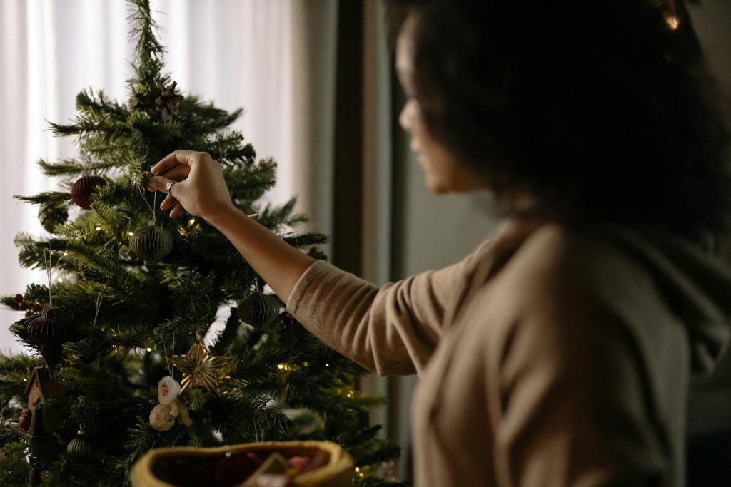 A woman decorates a Christmas tree indoors with ornaments, creating a festive atmosphere.