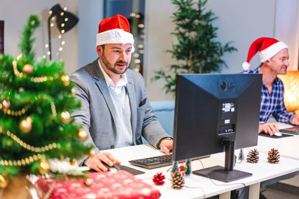 a man in a santa hat sitting at a computer with a man in a suit and a christmas