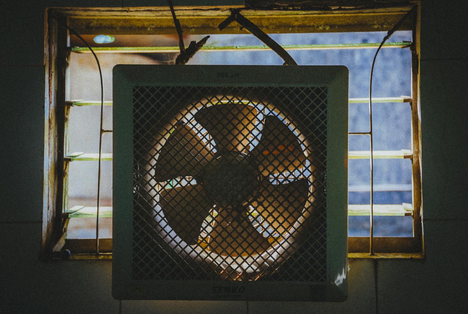A vintage exhaust fan installed in a rustic window frame, casting dramatic shadows.