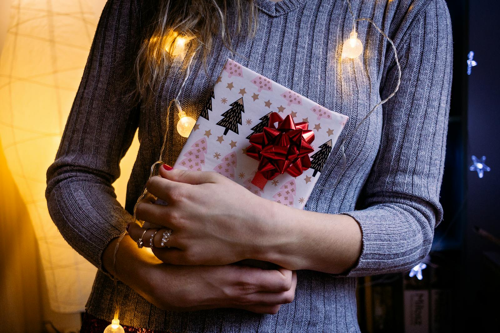 A woman holds a Christmas gift wrapped with a red bow and festive lights inside a cozy indoor setting.