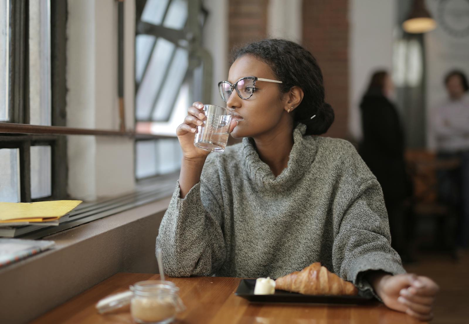 African American woman sipping water with a croissant at a cafe. Cozy indoor setting.