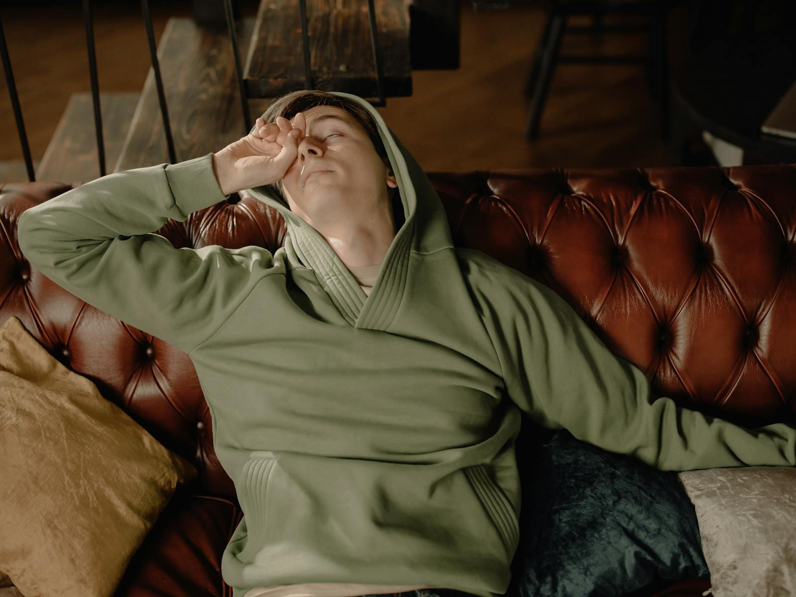A man in a green hoodie rests on a brown leather sofa, embodying a cozy and relaxed home atmosphere.