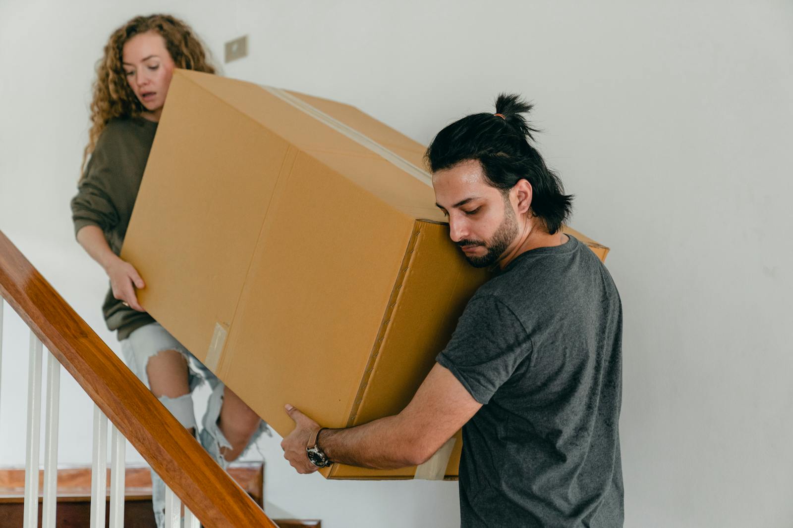 Focused couple in casual clothes carrying big carton box together down stairs in new house while moving personal items