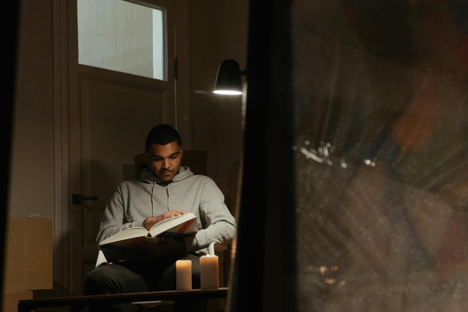 Man reading a book at home, under lamp light with candles, creating a warm and comfortable atmosphere.
