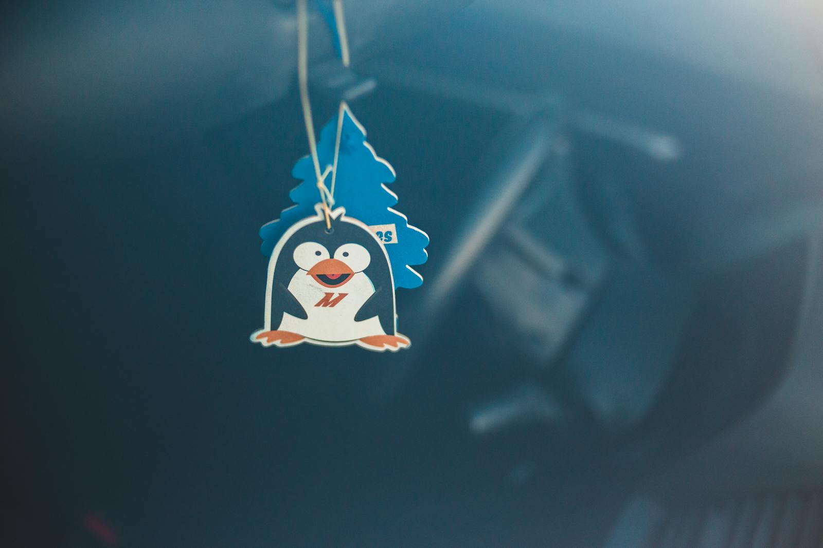 A cute penguin-themed air freshener hanging in a car with soft focus background.