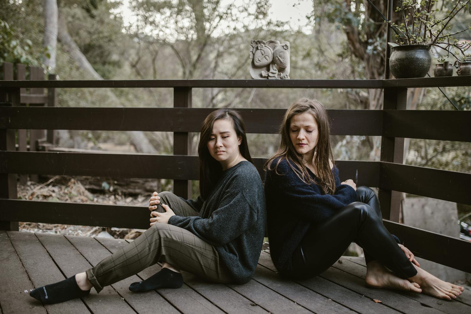 Two women sitting back to back on a wooden deck, deep in thought with nature surrounding them.