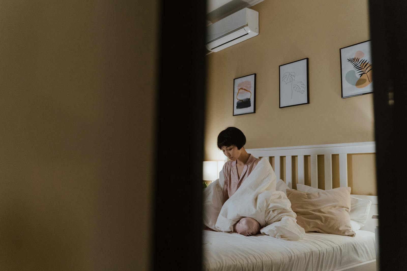 Asian woman sitting on bed, wrapped in blanket, indoors depicting feelings of loneliness.