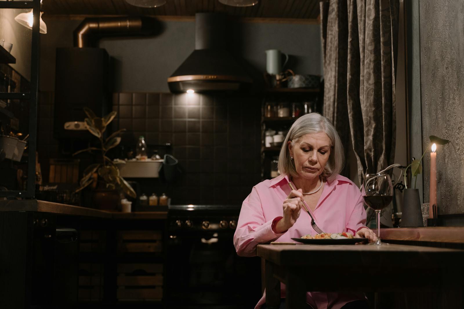 Elderly woman enjoying a solitary dinner by candlelight in a warm kitchen setting.