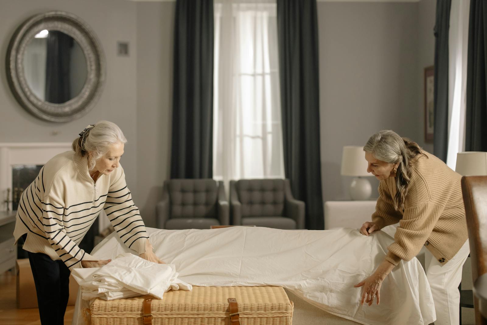 Two senior women folding sheets in a living room, preparing for moving out.