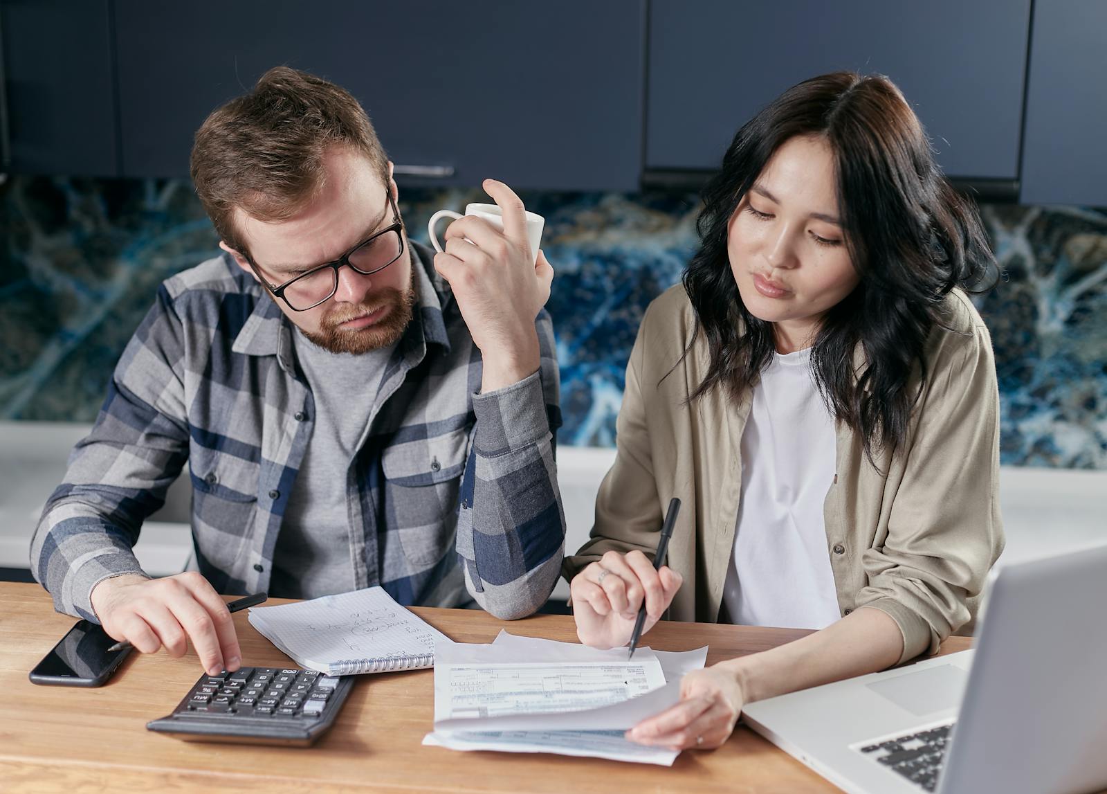 A couple reviewing household bills and budget using a calculator and laptop at their kitchen table.