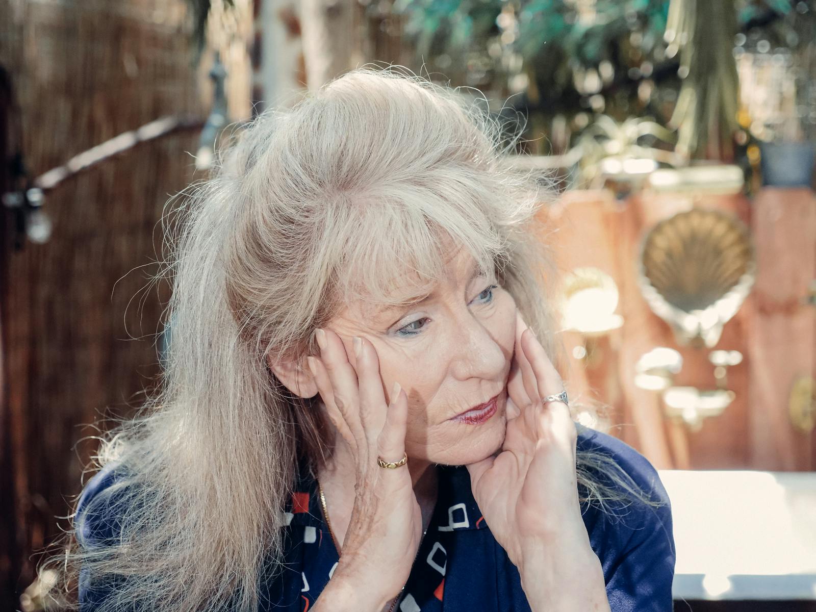 Thoughtful senior woman sitting outdoors, hands on face. A reflective moment captured in sunlight.