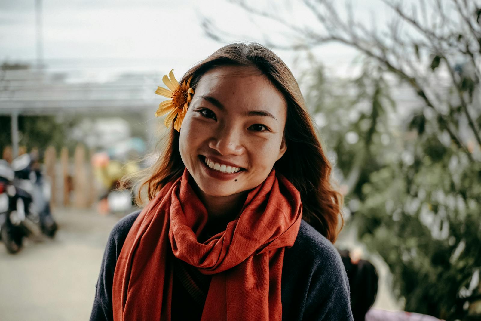 Warm portrait of an Asian woman smiling outdoors with a flower in her hair, wearing a scarf.
