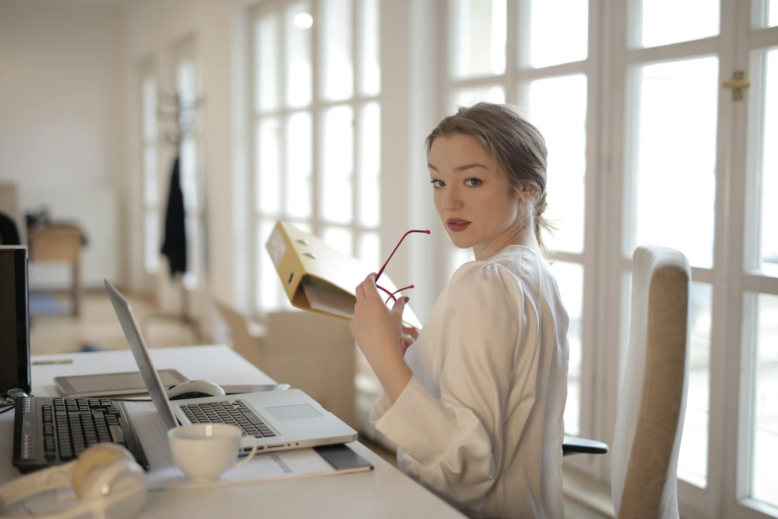 Elegant businesswoman sitting at desk, holding glasses, with a contemplative look.