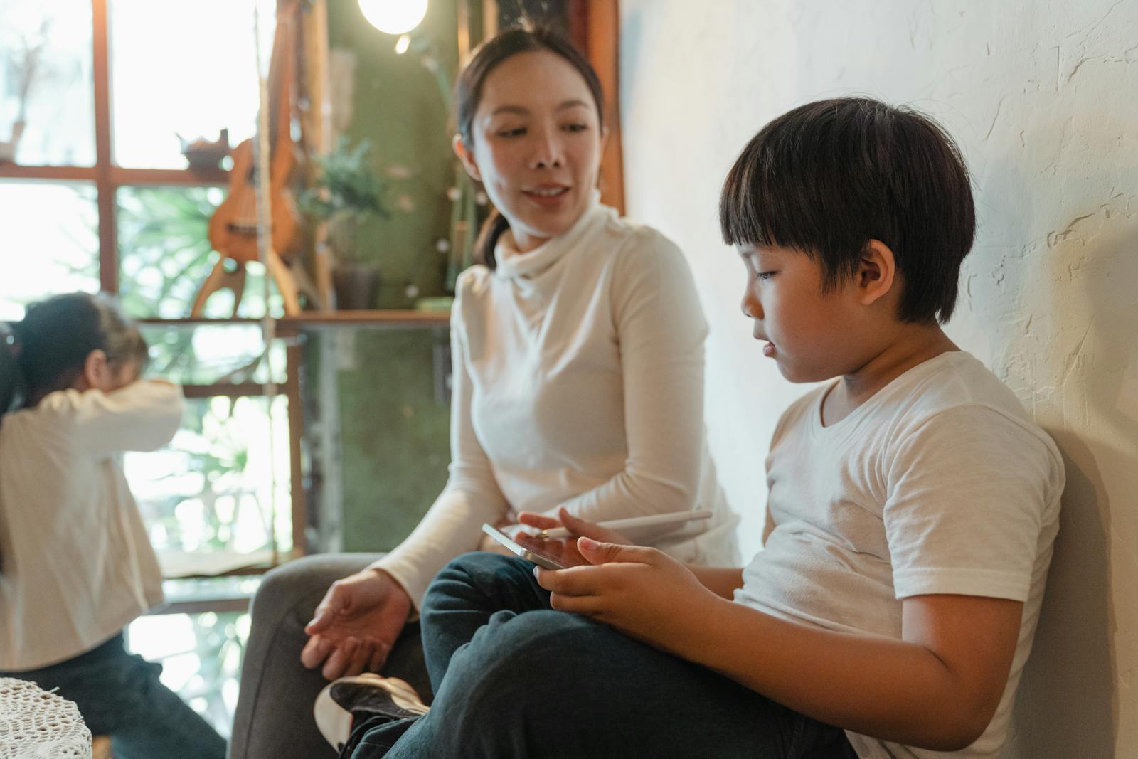 Positive ethnic woman sitting near cute son using tablet against adorable daughter looking at window while spending free time together at home